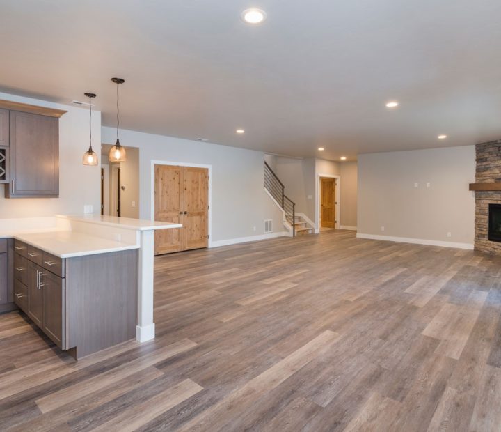 A spacious, modern basement with a kitchenette, light wood flooring, recessed lighting, a stone fireplace, and a staircase leading up, ideal for a multi-functional family space.