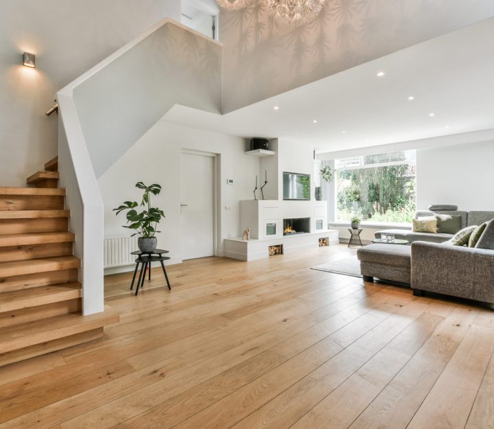 A contemporary living room with light wood flooring, a minimalist staircase, a gray sectional sofa, and large windows that highlight the clean lines and airy feel of the space.