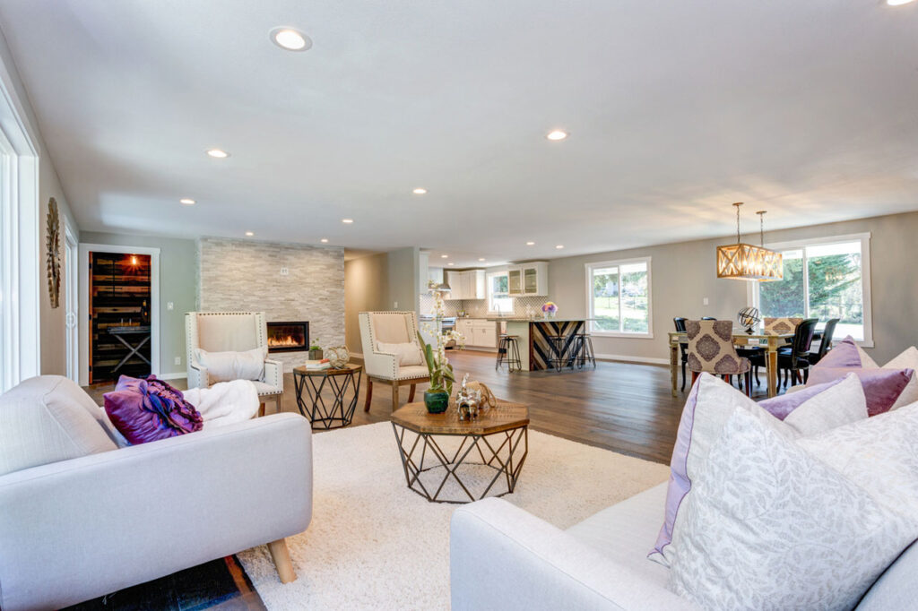 A beautifully remodeled open-concept living area featuring a stone fireplace, modern furniture, hardwood flooring, recessed lighting, and a seamless transition to the kitchen and dining space.