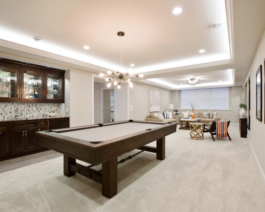 A spacious, finished basement featuring a wooden pool table, built-in bar with dark cabinets and mosaic backsplash, comfortable seating, and modern lighting for entertainment.